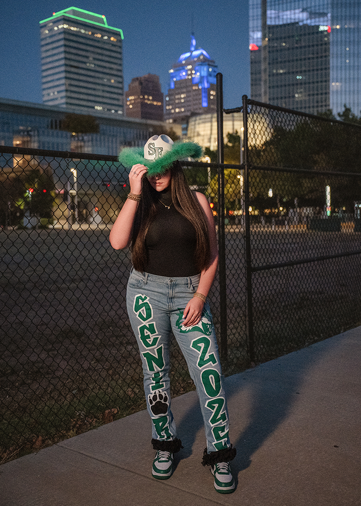 A high school senior poses confidently at dusk by a fence, wearing a fluffy hat and custom jeans labeled "Senior 2025." Oklahoma City skyscrapers glow in the background.