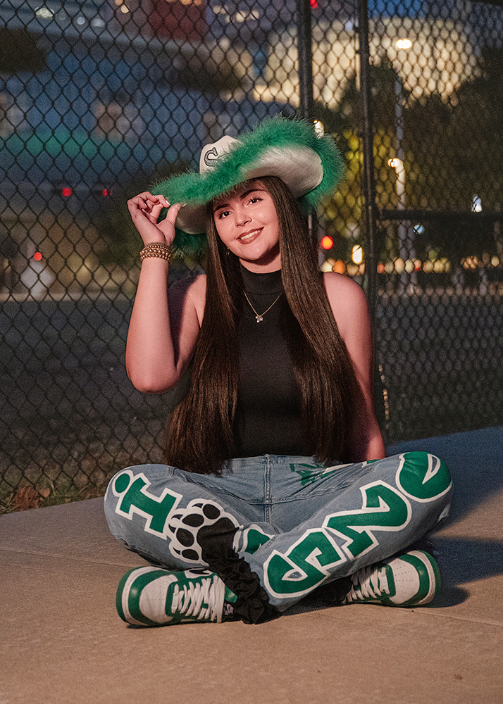 A high school senior sits cross-legged on concrete, wearing a hat with green trim, a black sleeveless top, and jeans with bold green lettering. A chain-link fence is in the background, with city lights softly glowing, creating a relaxed, urban vibe.