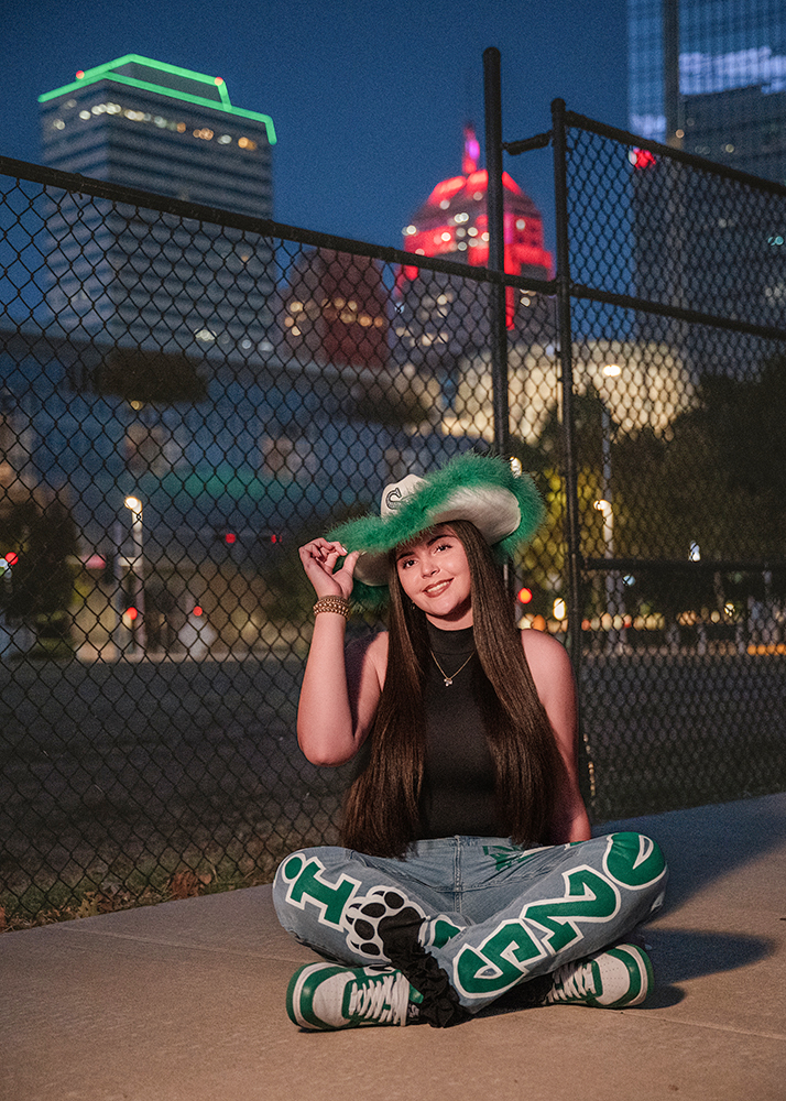 Senior girls sits cross-legged on a sidewalk at dusk, wearing a green cowboy hat and bold patterned pants. Cityscape with lit buildings in the background.