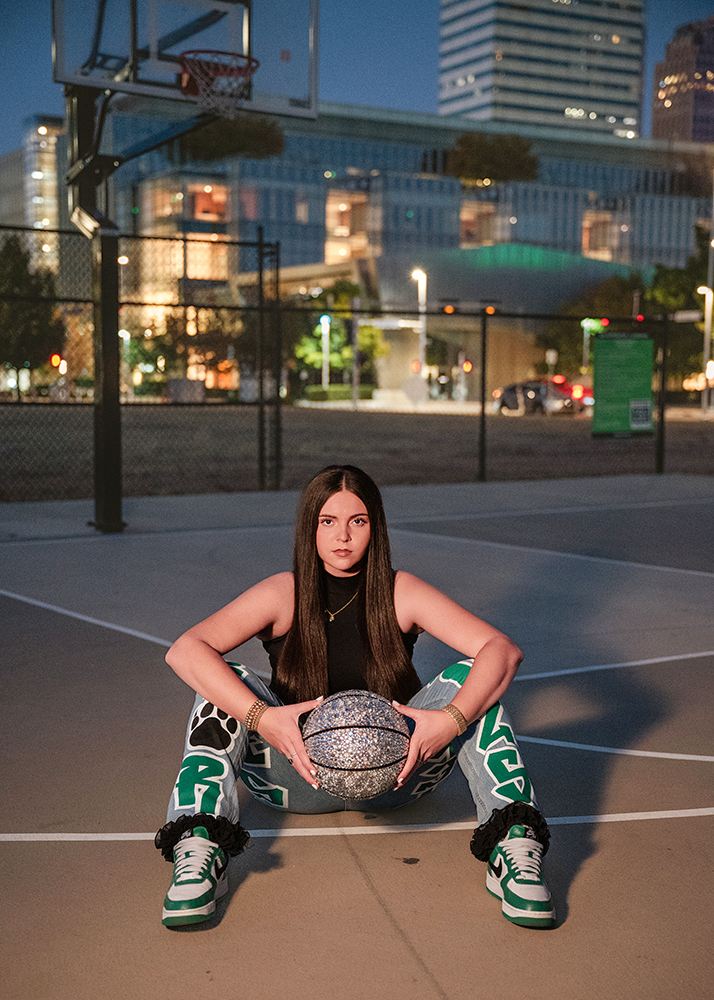 Senior girl in urban basketball court at dusk holds a sparkling basketball, wearing a black top and colorful pants. Background shows city lights.