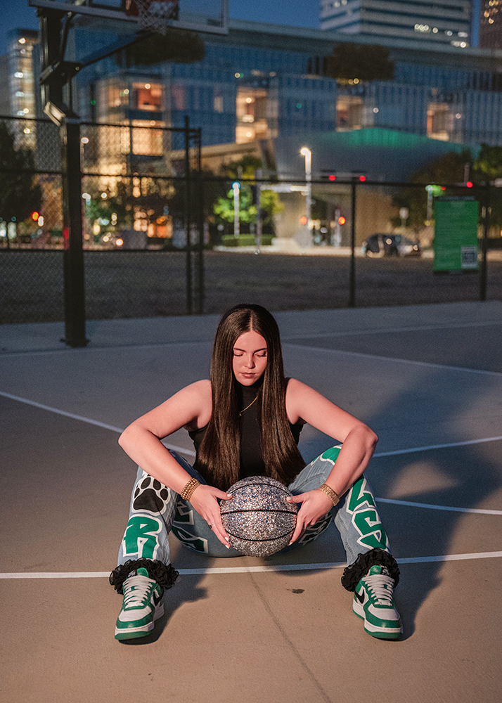 A high school senior with long hair sits on an outdoor basketball court at dusk, holding a sparkling basketball. She wears colorful sneakers and patterned pants, conveying a contemplative mood.