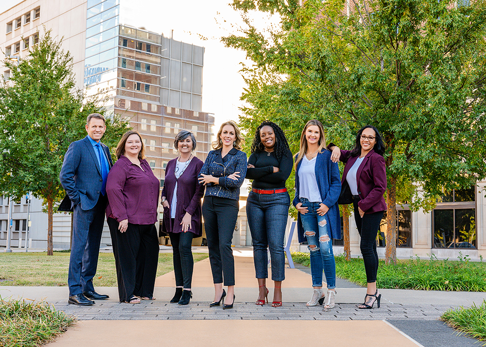 A group of seven professionally dressed individuals stand confidently in an urban park. Tall buildings and greenery surround them.