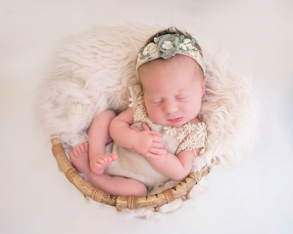 A peaceful newborn, wearing a floral headband and lace outfit, sleeps in a wicker basket lined with soft, white fur, conveying warmth and serenity.