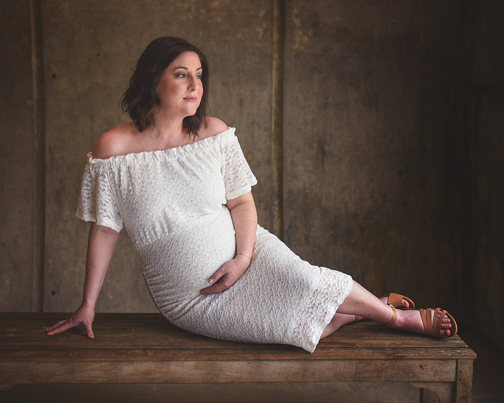 A pregnant woman in a white, off-the-shoulder dress reclines on a wooden bench, her hand on her belly.