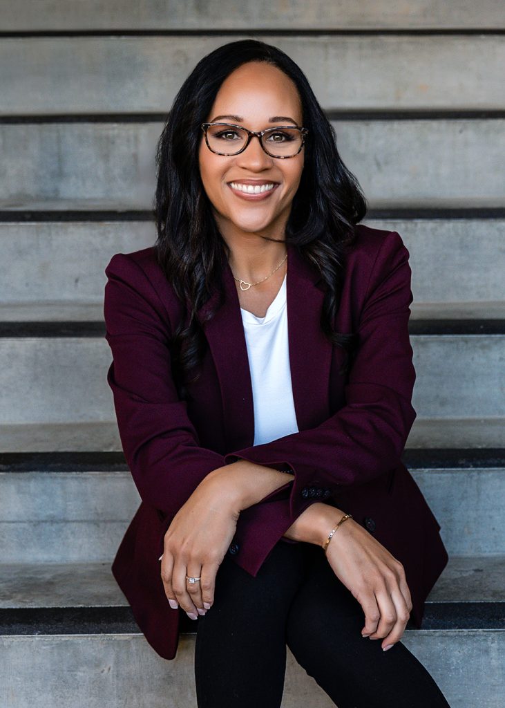 Smiling person seated on stairs wearing glasses and a maroon blazer. Arms are crossed on knees, conveying confidence and warmth.