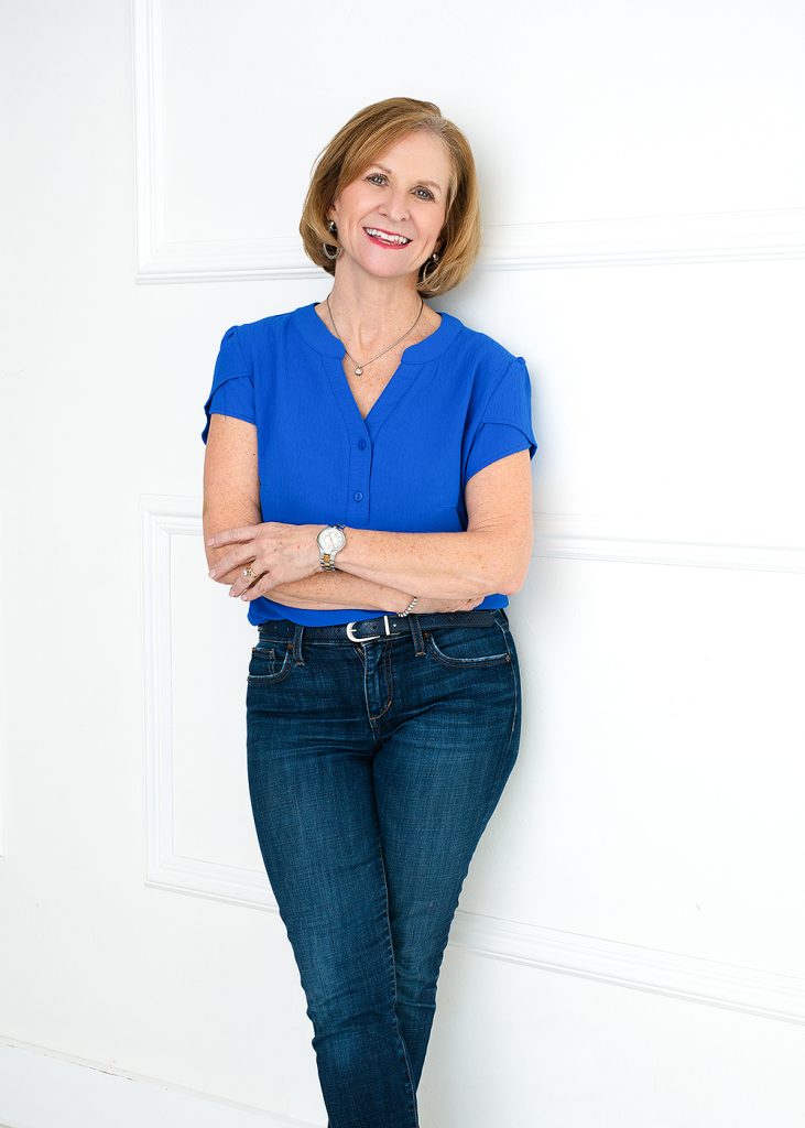 A smiling woman in a blue blouse and jeans stands with arms crossed against a white wall. Her relaxed posture and warm expression convey friendliness.