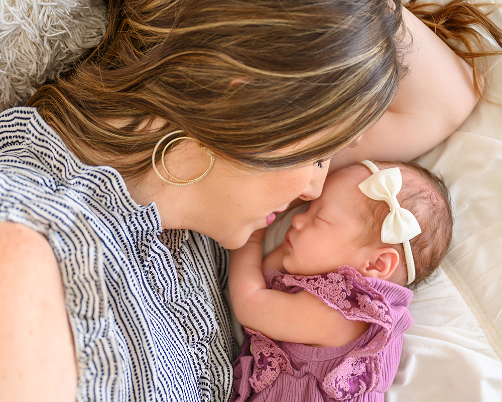 A mother gently nuzzles her newborn, both resting on a soft pillow. The baby wears a purple dress and a white bow headband