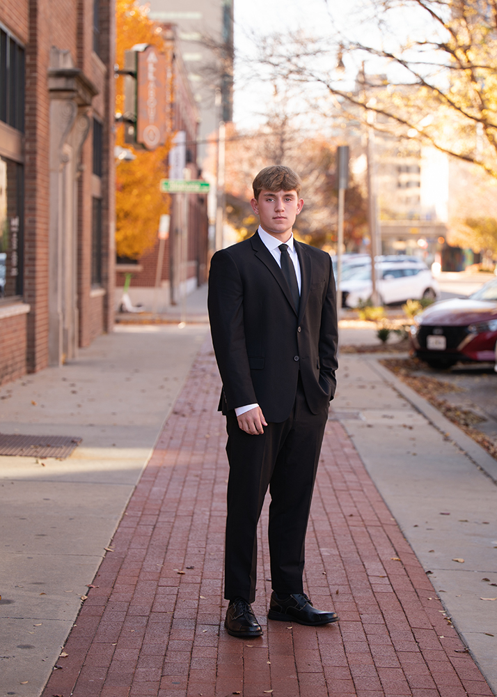 A senior boy in a black suit stands on a sunlit urban sidewalk in Bricktown in OKC with autumn leaves around. The scene is calm, with a backdrop of brick buildings.