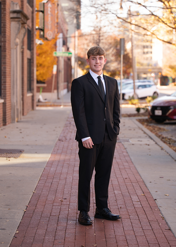 A high school senior in a black suit and tie stands confidently on a sunlit brick sidewalk. Autumn leaves and city buildings create a warm urban backdrop.