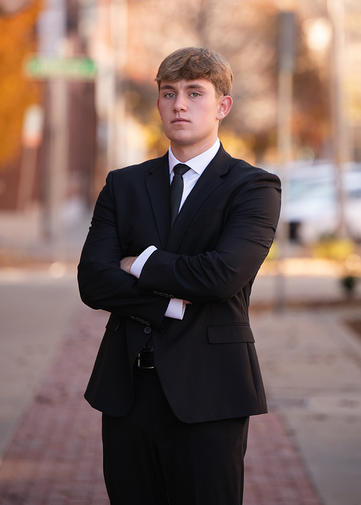 A senior guy in a black suit with a white shirt and black tie stands confidently with crossed arms on a street. Autumn foliage in the blurred background.