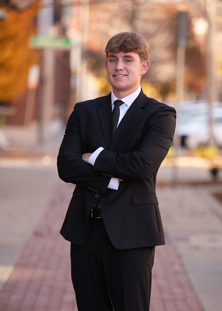 A high school senior in a black suit and tie stands confidently with arms crossed on a city sidewalk. The background is softly blurred with autumn colors.