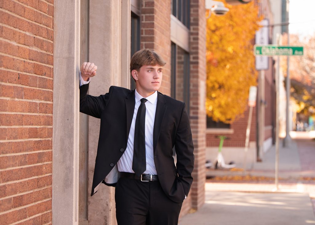 Young man in a suit leans against a brick wall on a sunny day. He looks thoughtful. Vibrant autumn trees and street signs in the background.