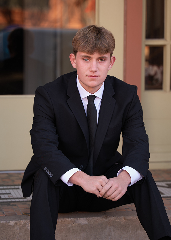 Senior boy in a black suit and tie sits on stone steps, looking confidently at the camera. The neutral background adds a professional tone.
