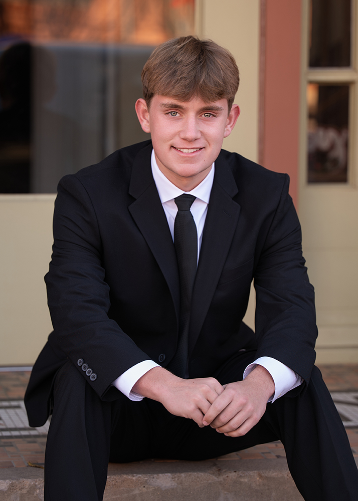 High school senior wearing a black suit and tie, sitting on steps, smiling softly. The background is a blurred doorway and wall, creating a formal, warm tone.