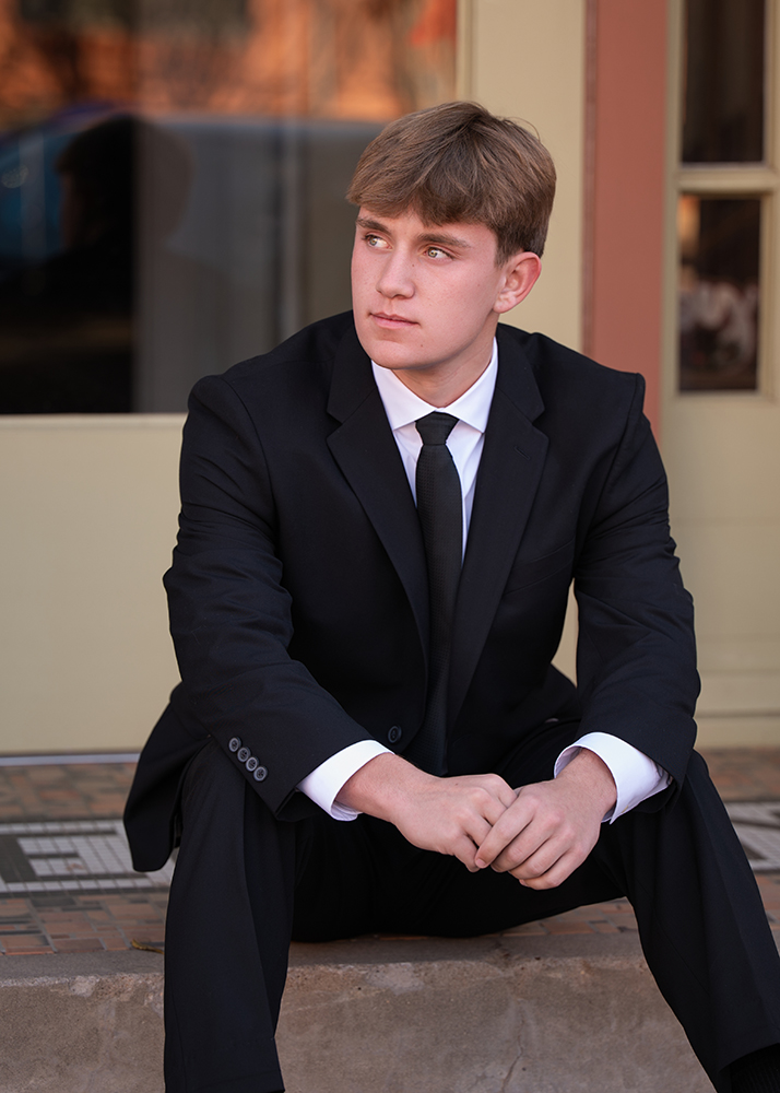 Young man in a black suit and tie sits thoughtfully on a step. He looks to the side with a calm expression. Warm, casual setting in the background.