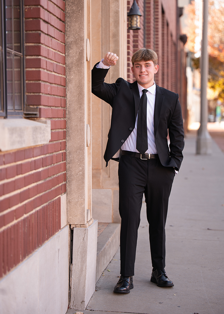 Senior session for William in a black suit leans casually against a brick building in OKC on a sunny day. He smiles confidently, with a hand in his pocket.
