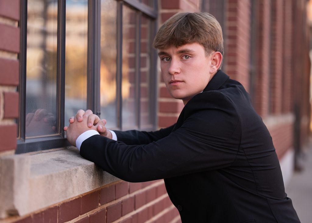 High school senior in a black suit leans on a brick wall, gazing thoughtfully out a window. The setting has a calm, contemplative tone with soft lighting.