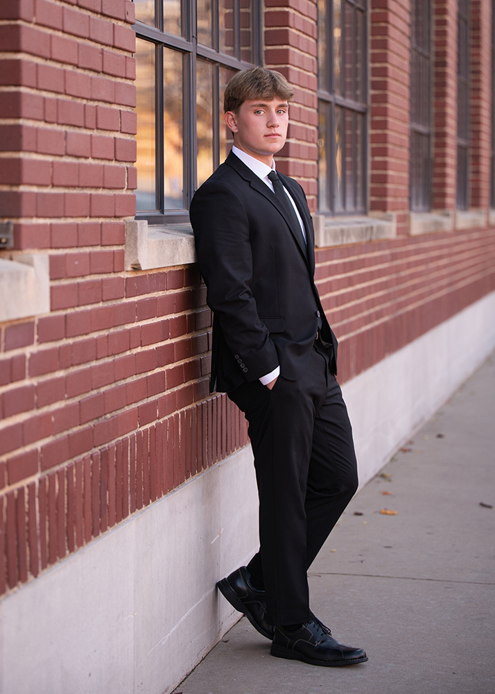Senior guy in a black suit and tie leans confidently against a brick building wall. The setting feels formal, with a calm and composed atmosphere.