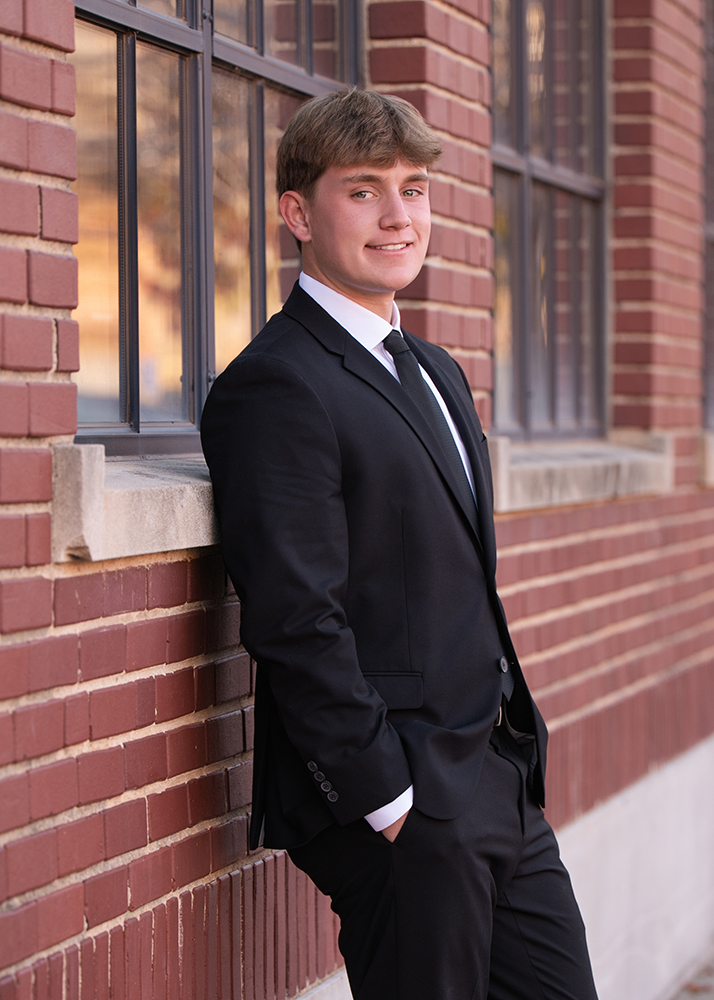 High school senior in a black suit and tie leans confidently against a brick building with hands in pockets, smiling, under bright natural light.