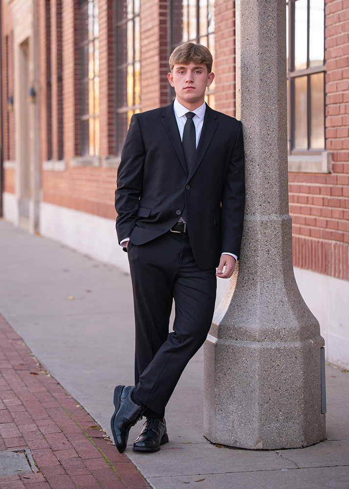 High school senior in a black suit leans against a concrete pillar on a sidewalk beside a brick building. He looks confident and composed.