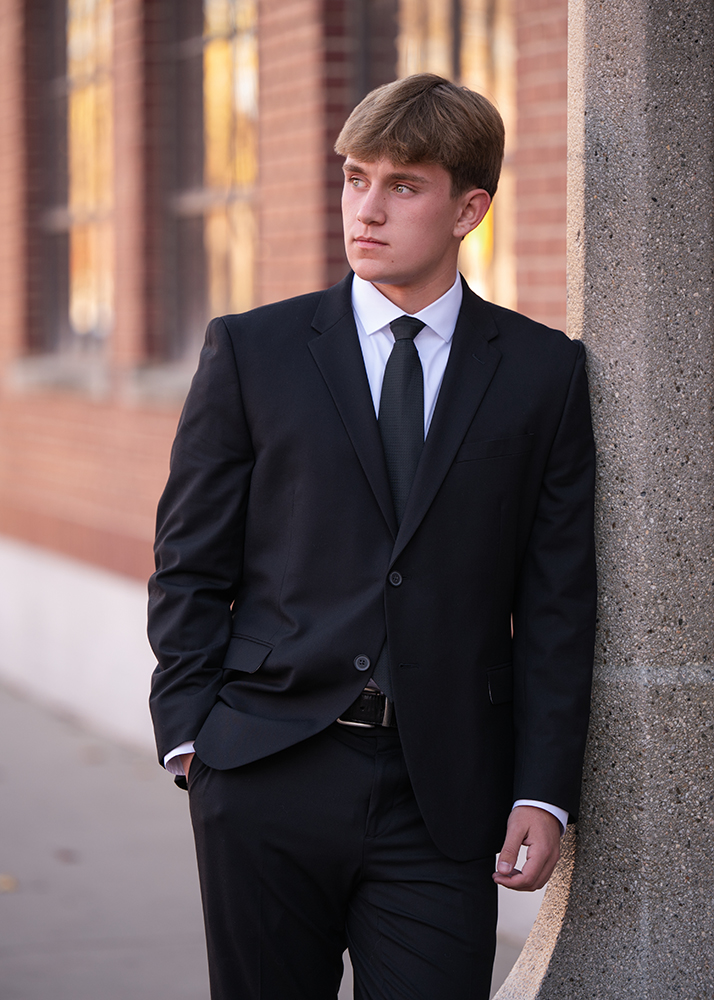 A young man in a black suit and tie leans against a column, gazing to the side. The background features a brick wall with windows, creating a professional and contemplative tone.