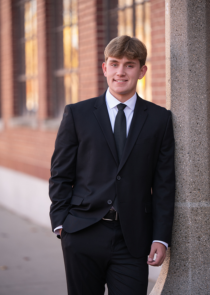 Senior boy in a black suit and tie leans against a stone pillar, smiling. The background features a brick building with tall windows, conveying a professional tone.