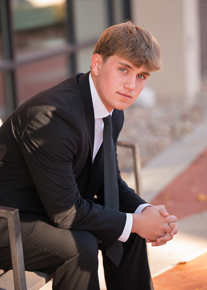 Senior photos of a young man in a black suit sits thoughtfully on a bench, hands clasped. He's outside a modern building, with soft lighting creating a calm mood.