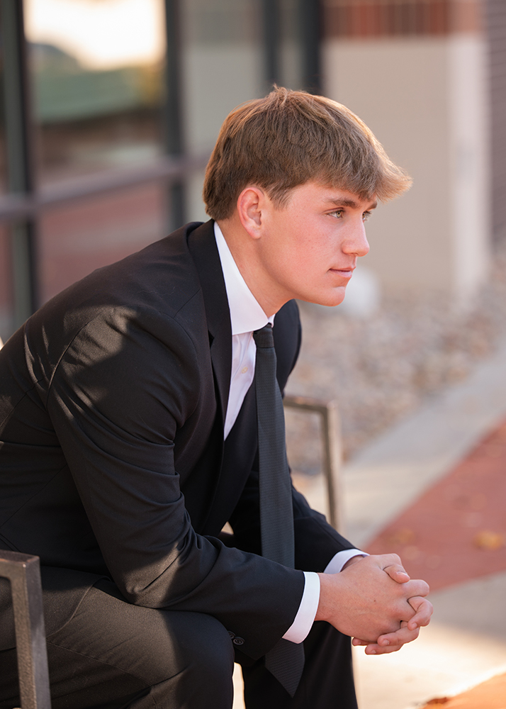 High school senior photos of a young man in a black suit and tie sits on a bench, hands clasped, looking contemplative. The background features blurred building windows.