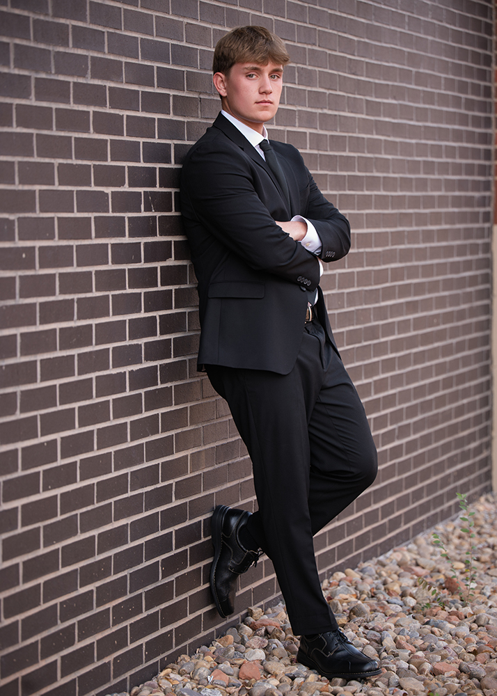 A high school senior in a black suit and tie leans against a brick wall with arms crossed, conveying a confident and serious demeanor. Rocky ground below.