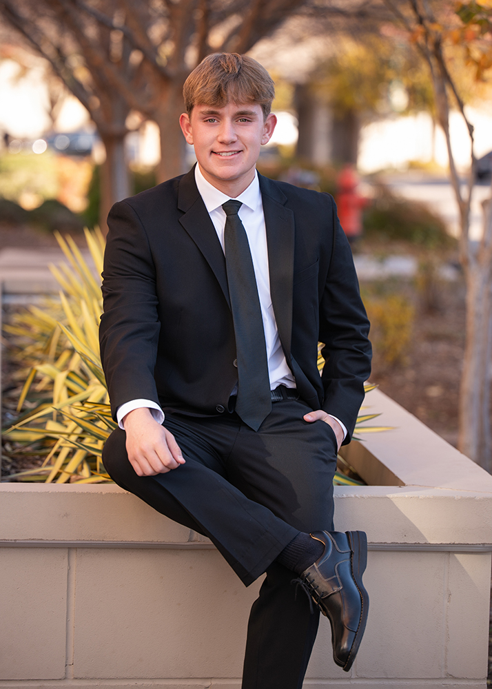 Senior boy in a suit sits on a ledge outdoors, smiling confidently. He wears a black suit and tie. The background features trees and soft, natural light.