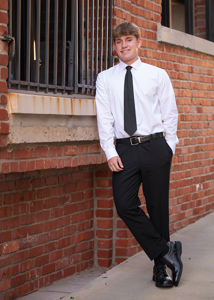 Senior guy in a white shirt and black tie leans casually against a red brick wall, conveying a confident and relaxed attitude.