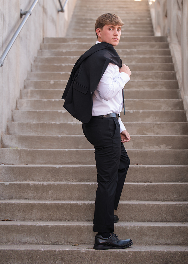Young man taking his senior photos in business attire stands on concrete steps, looking back over his shoulder. He carries a black jacket over one shoulder, exuding confidence.