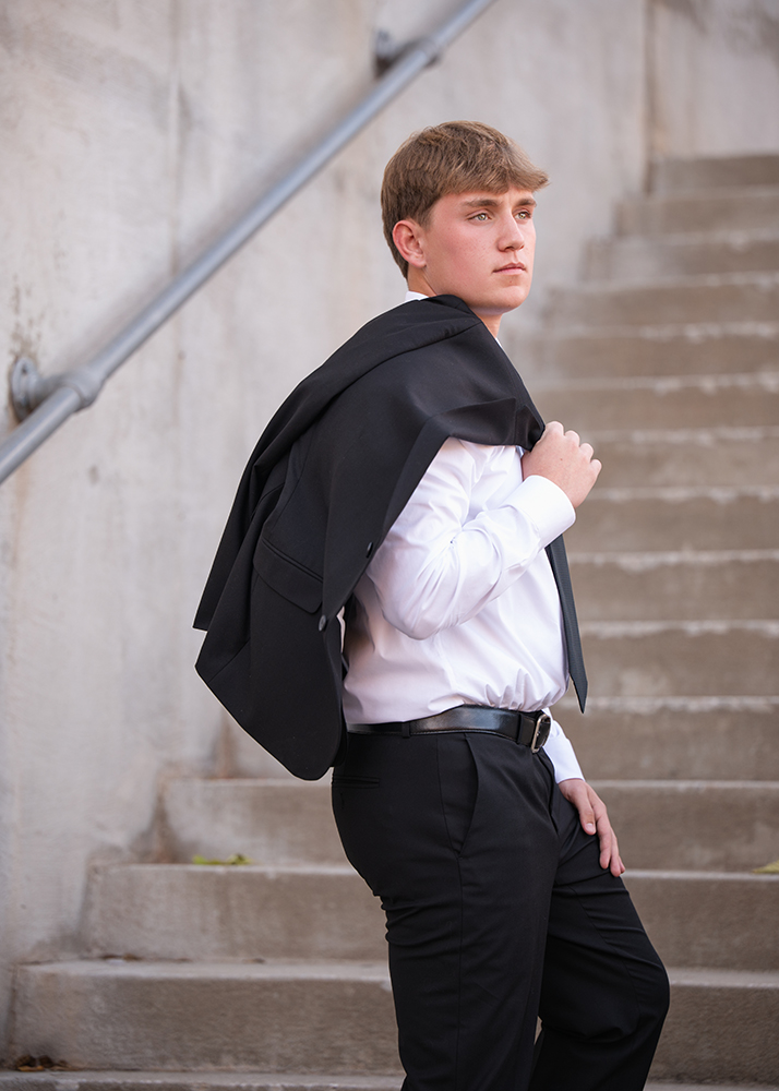 Young man taking his senior photos in business attire stands on concrete steps, looking back over his shoulder. He carries a black jacket over one shoulder, exuding confidence.