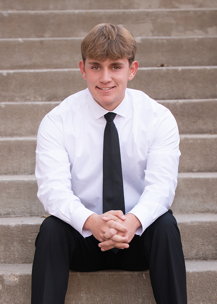 Young man seated on concrete steps, wearing a white dress shirt, black tie, and black pants. He smiles confidently, conveying a formal tone.