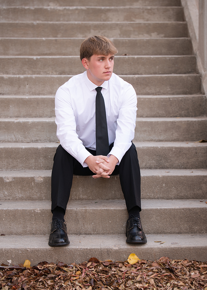 High school senior in a white shirt and black tie sits pensively on outdoor steps, hands clasped. Fallen leaves scatter below, suggesting a contemplative autumn setting.