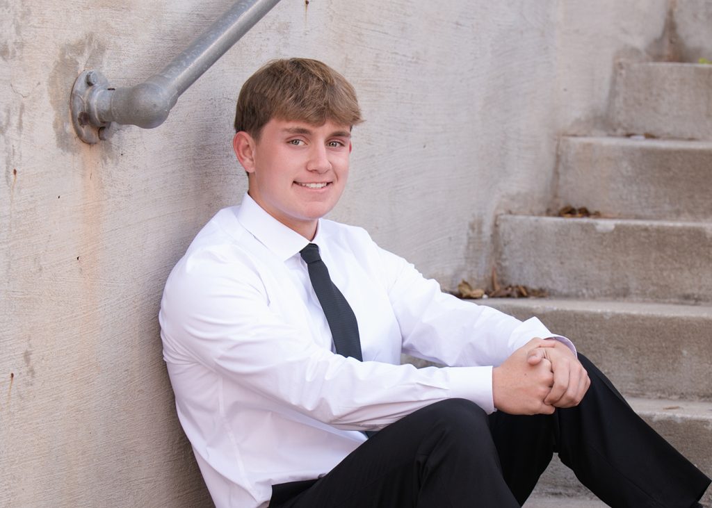 High school senior in a white shirt and black tie sits on concrete steps, smiling, leaning against a wall in Bricktown Oklahoma City. The scene is calm and casual.