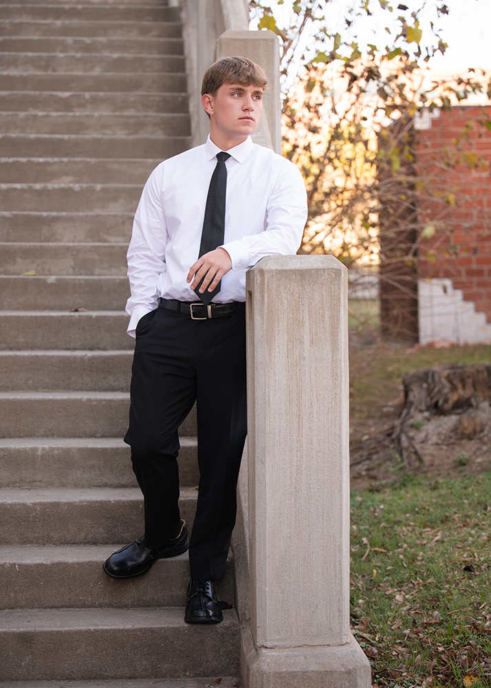 Senior guy in a white shirt and black tie stands on outdoor steps, leaning against a pillar. He looks thoughtfully into the distance. The scene has an autumnal feel.