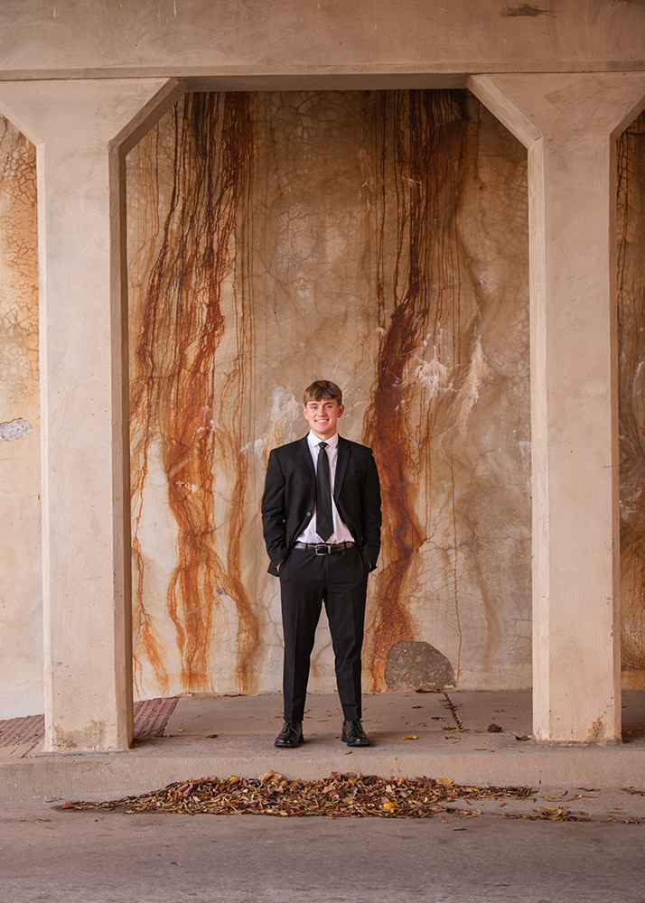 High school senior in a black suit and tie stands under a concrete structure with rust-streaked walls. Leaves scattered on the ground evoke an autumn feel.