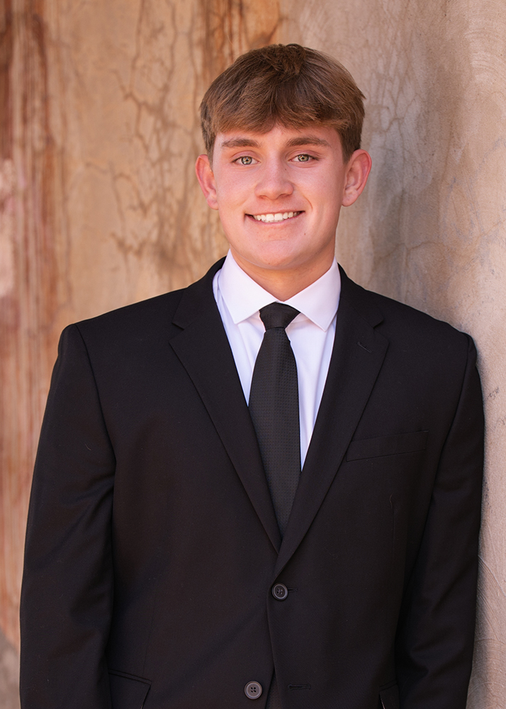 Senior boy in a black suit and tie smiles warmly against a textured, earth-toned wall. The atmosphere is professional yet approachable.