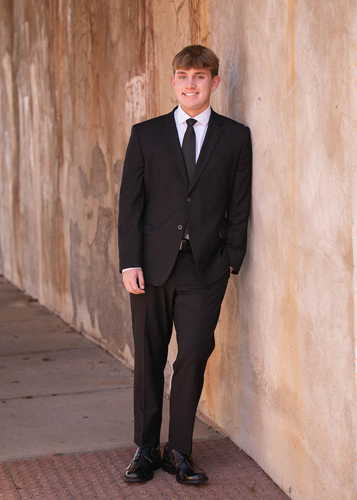 A senior boy in a black suit and tie leans against a textured wall, smiling warmly. The setting is an urban sidewalk. Professional and confident vibe.