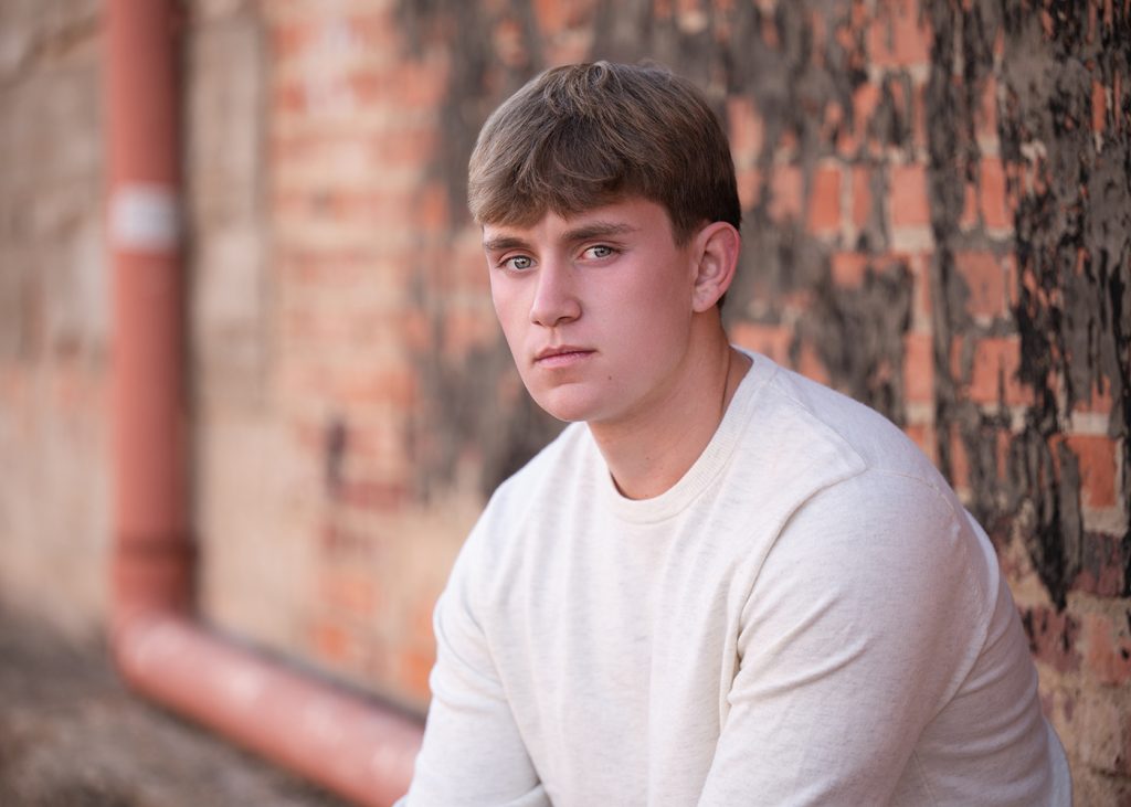 A high school senior in a white sweater sits against a weathered brick wall. The atmosphere is contemplative, with muted colors and soft lighting.