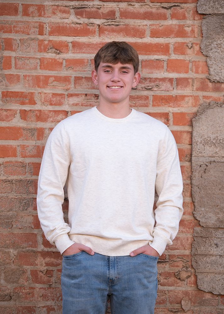 Smiling high school senior in a light sweatshirt and jeans stands against a textured brick wall, hands in pockets, conveying a relaxed and casual vibe.