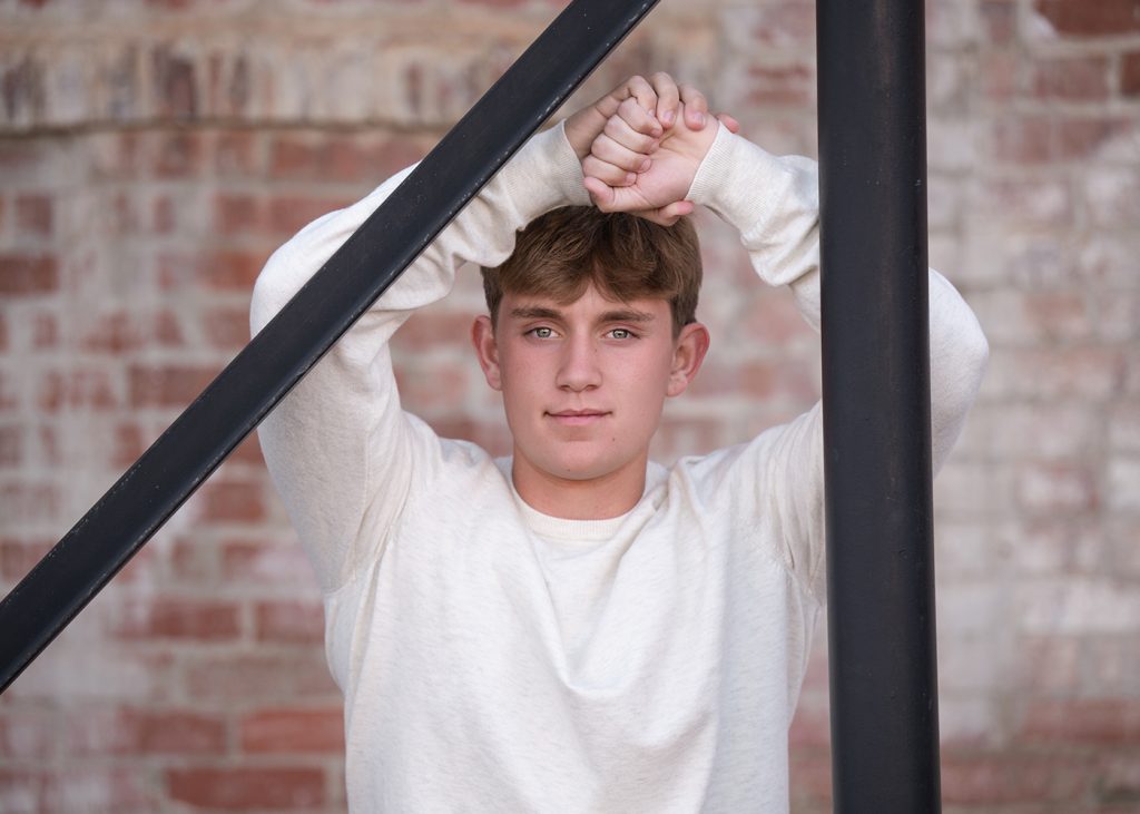 Senior guy in a white sweater leans against a black metal beam, hands clasped above his head. He stands against a brick wall with a calm expression.