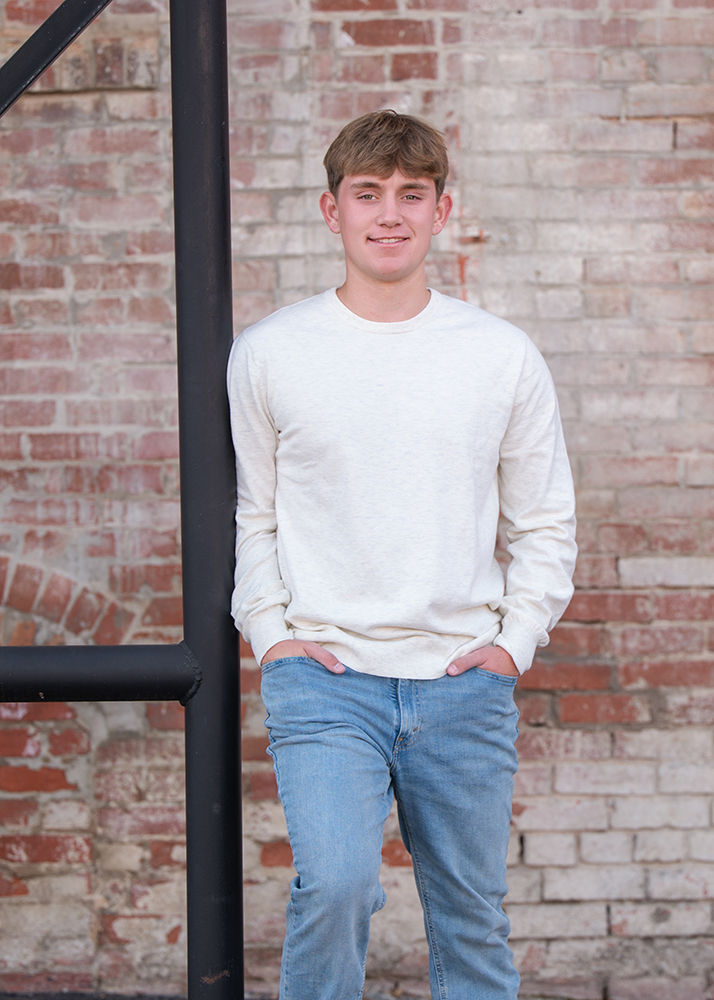 Senior boy in a white sweater and jeans leans against a metal beam, hands in pockets, in front of a rustic brick wall, exuding a relaxed demeanor.