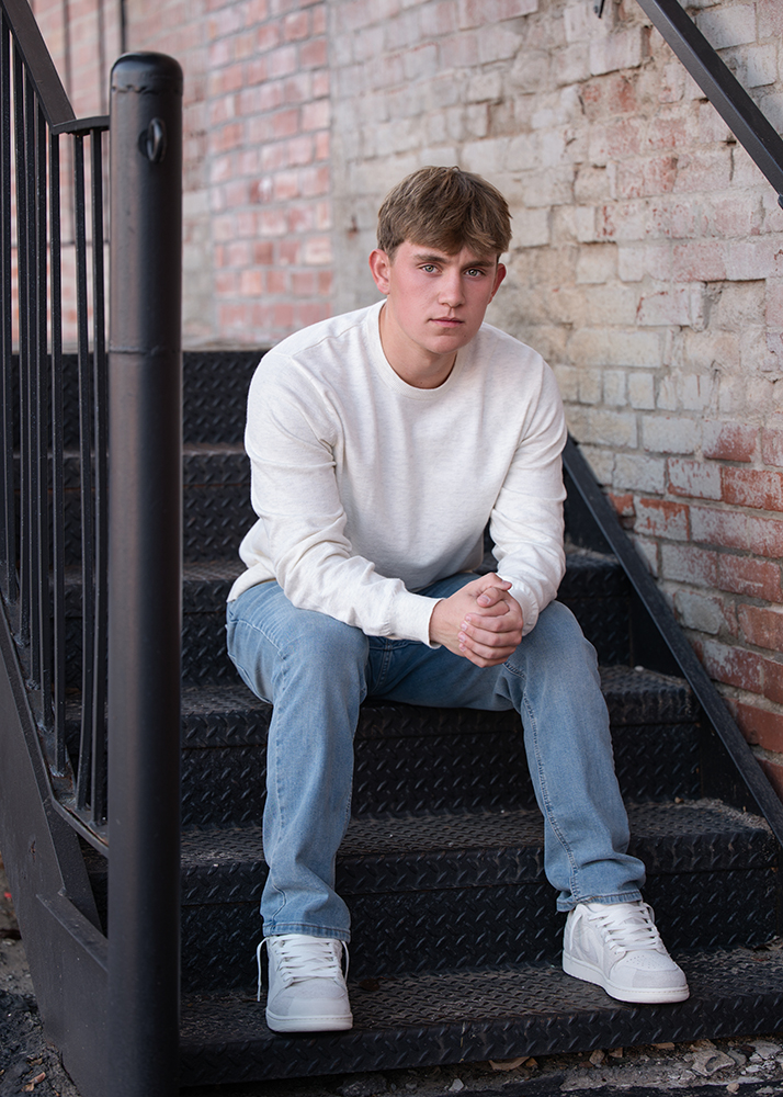 High school senior in a white sweater and jeans sits on metal stairs, framed by a brick wall. He gazes pensively, conveying a contemplative mood.