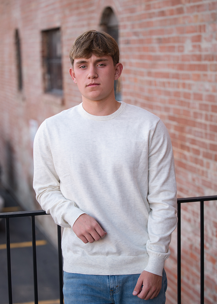 High school senior in a white sweater and blue jeans leans against a black railing, set in front of a brick wall. He appears calm and composed.