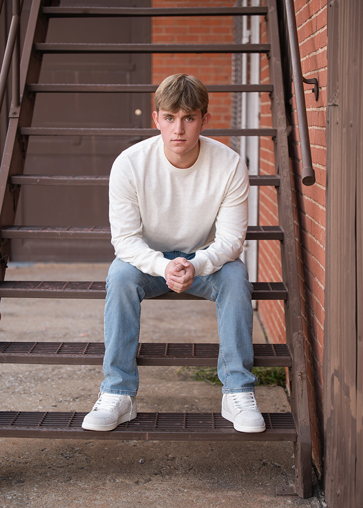 Senior boy sits on metal stairs against brick wall, wearing a white sweater, blue jeans, and white sneakers, with a focused expression.