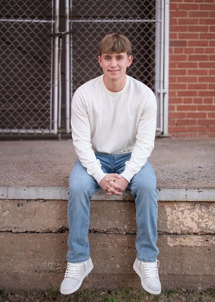 Photos of a senior guy sitting on a concrete step in front of a metal fence. They're wearing a white sweater, blue jeans, and white sneakers, smiling gently.