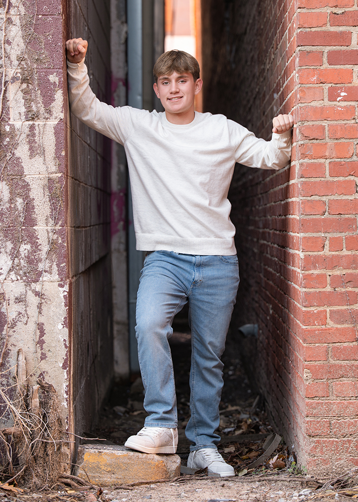 High school senior in a white sweater and jeans stands in a narrow alley with brick and weathered walls, smiling confidently while leaning on the walls.