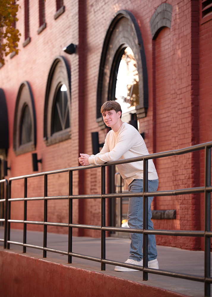 A high school senior in a white sweater and jeans leans on a metal railing, smiling. The background features a red brick building in downtown OKC with arched windows, creating a warm, relaxed atmosphere.
